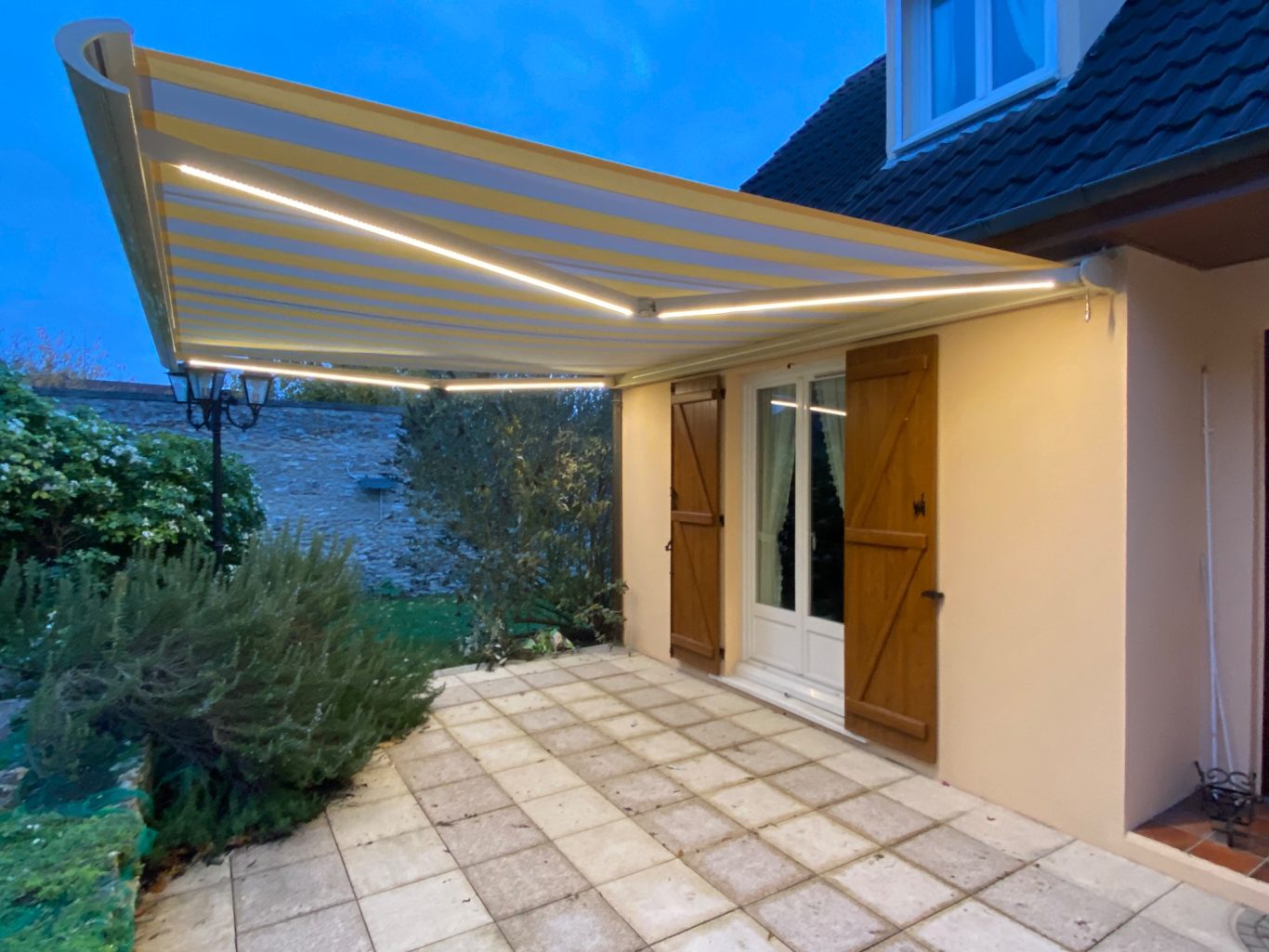 Auvent déplié sur une terrasse, illuminé, avec des portes ouvertes et des plantes autour.