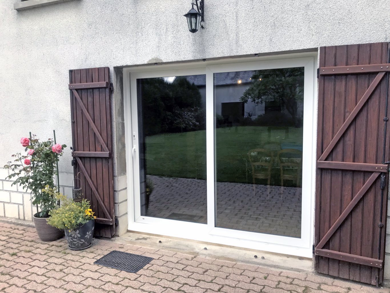 Porte-fenêtre avec volets en bois et fleurs devant, donnant sur un jardin verdoyant.