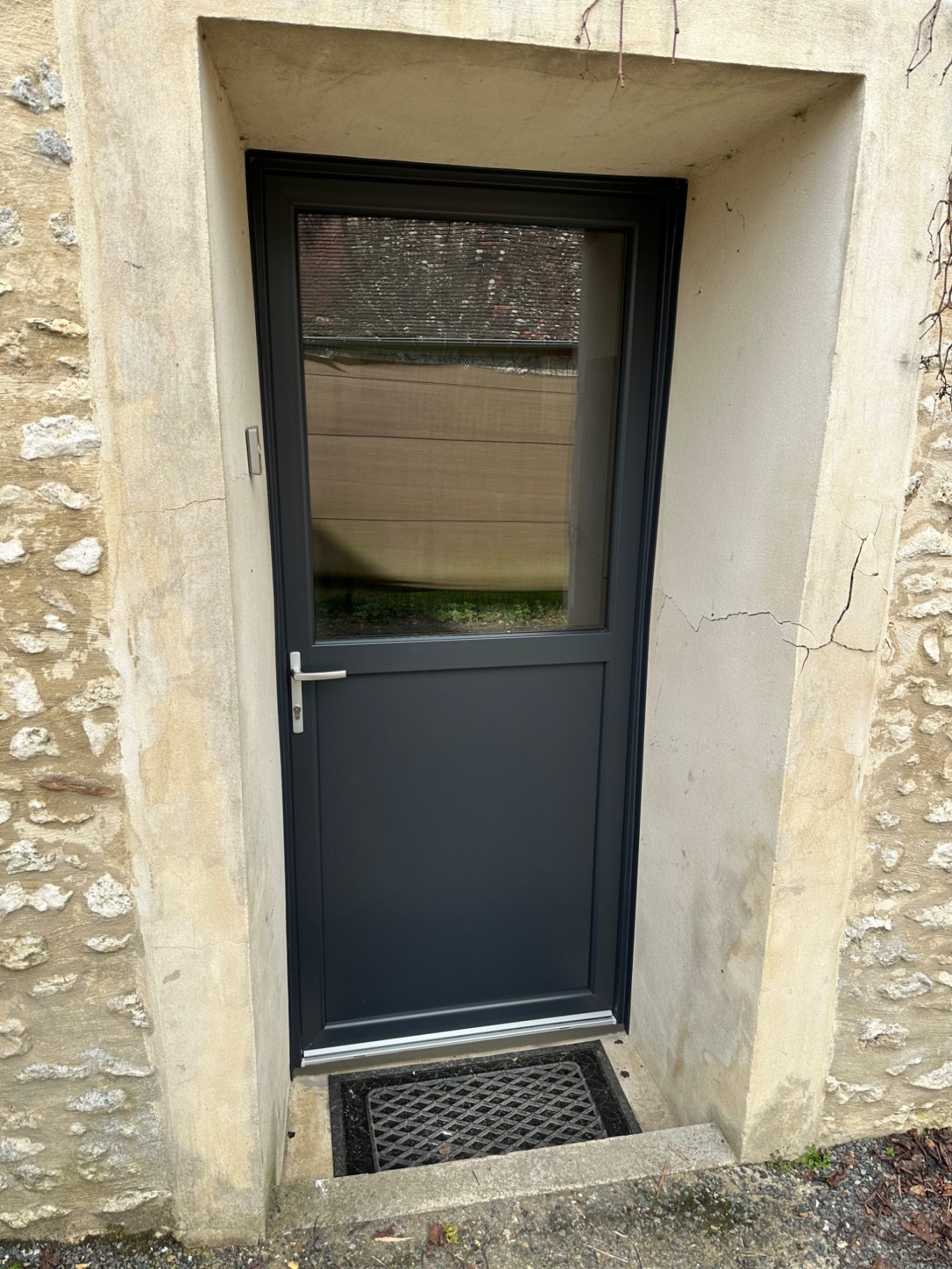 Porte en métal gris avec une fenêtre, encadrée par un mur en pierres.