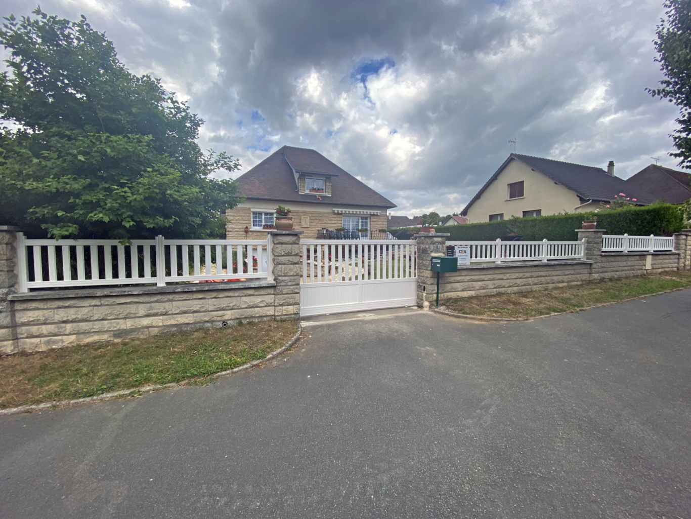 Maison avec clôture blanche, portail fermé et ciel nuageux. Rue calme à proximité.