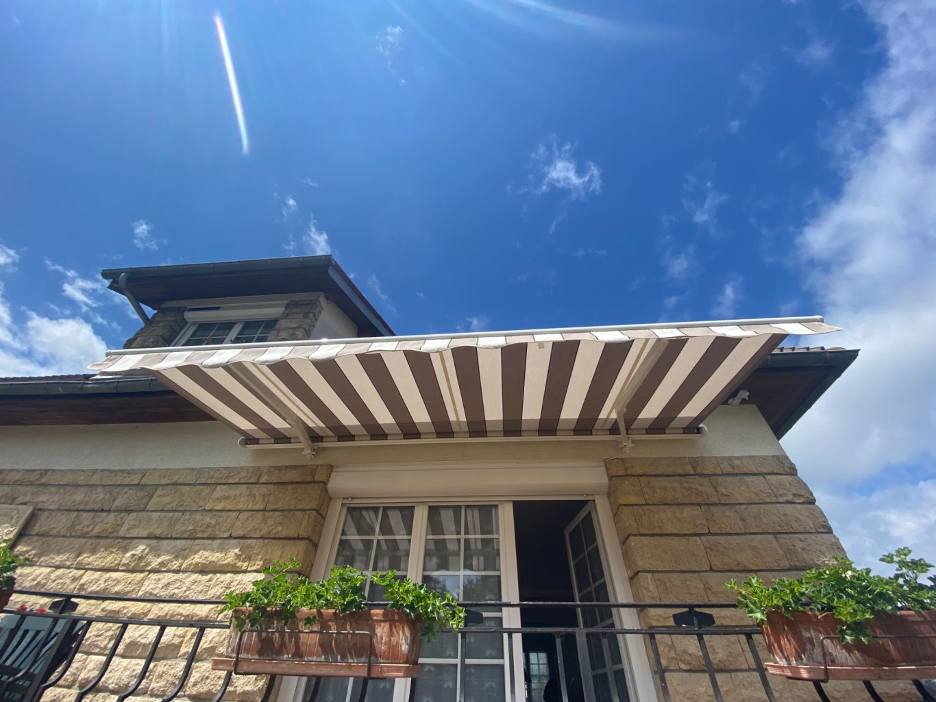 Maison en pierre avec un auvent rayé et des plantes sur le balcon, sous un ciel bleu.