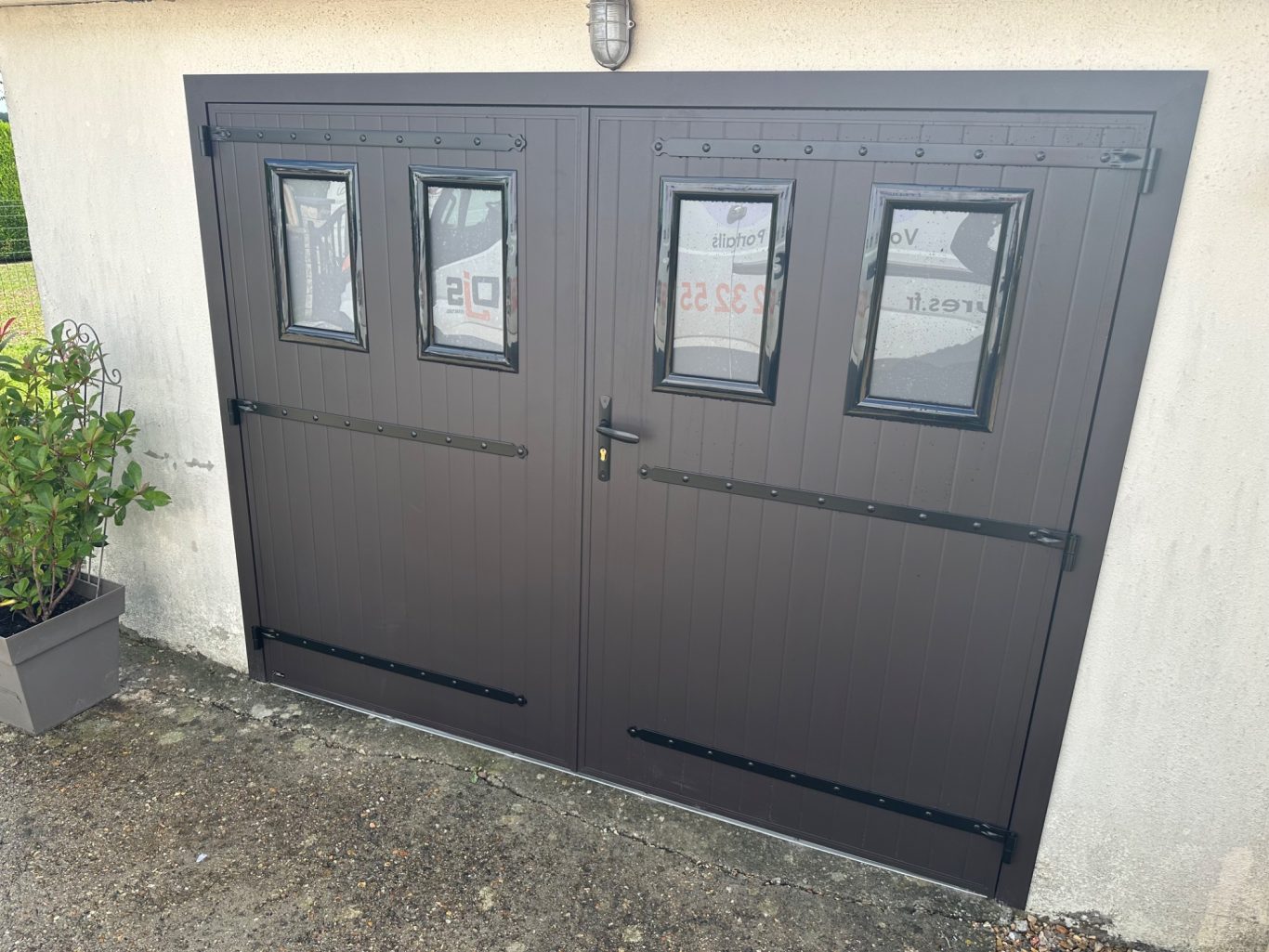 Portail de garage en métal marron, avec quatre fenêtres en verre. Plante à côté.