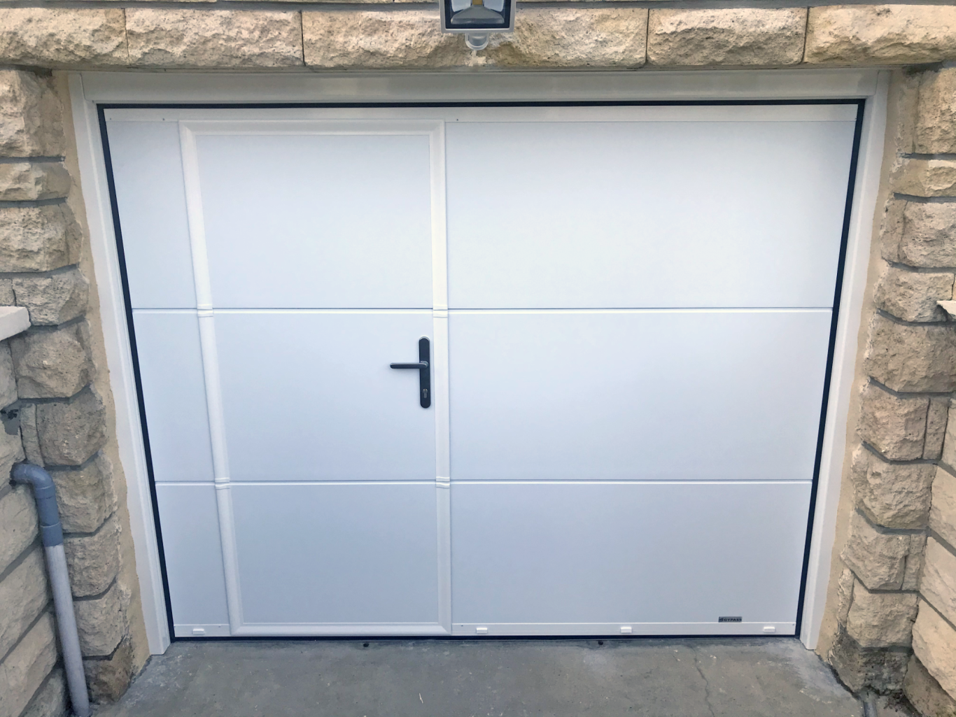 Porte de garage blanche avec poignée, encadrée de pierres claires.