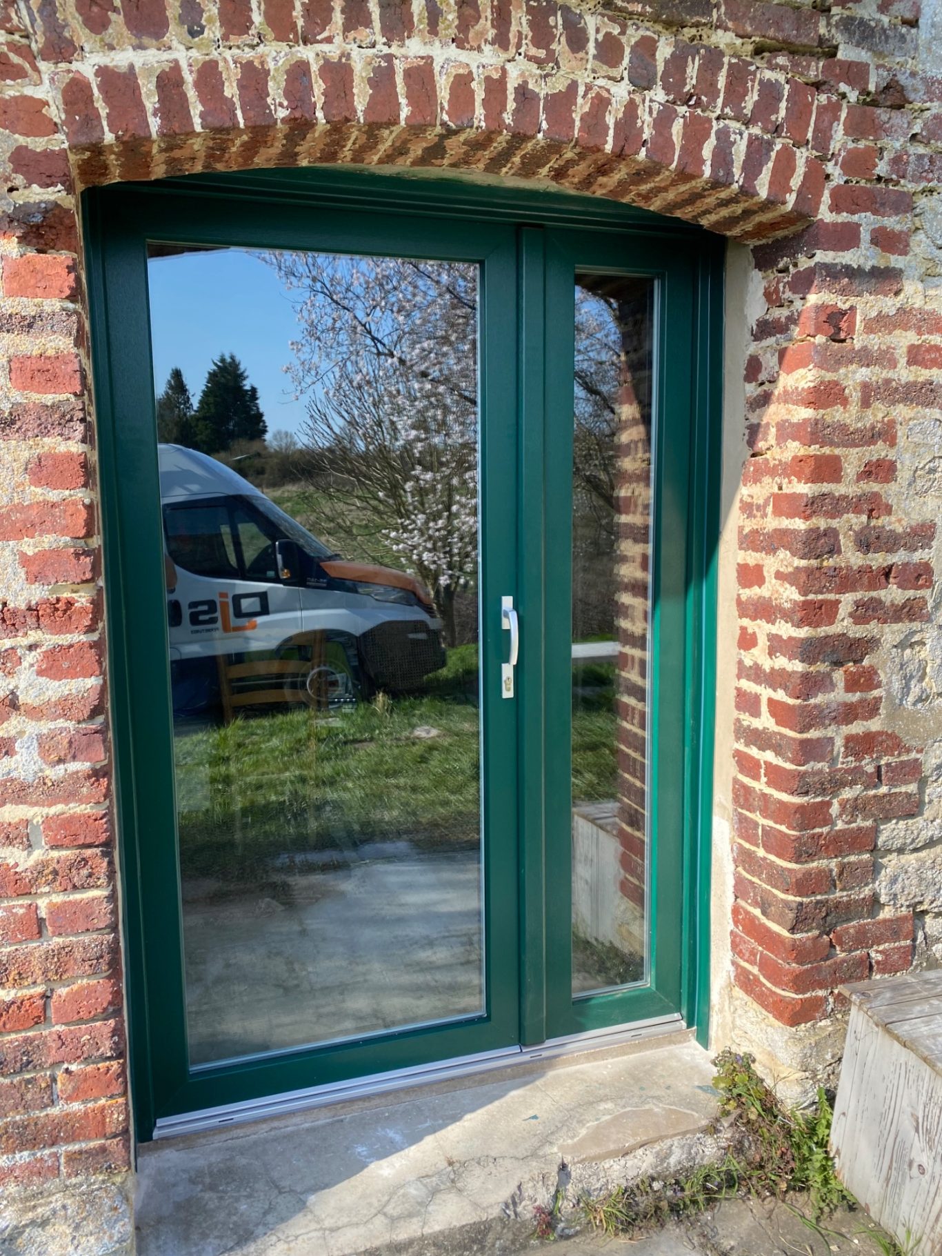 Porte vitrée verte encadrée de briques, ouverte sur un jardin.