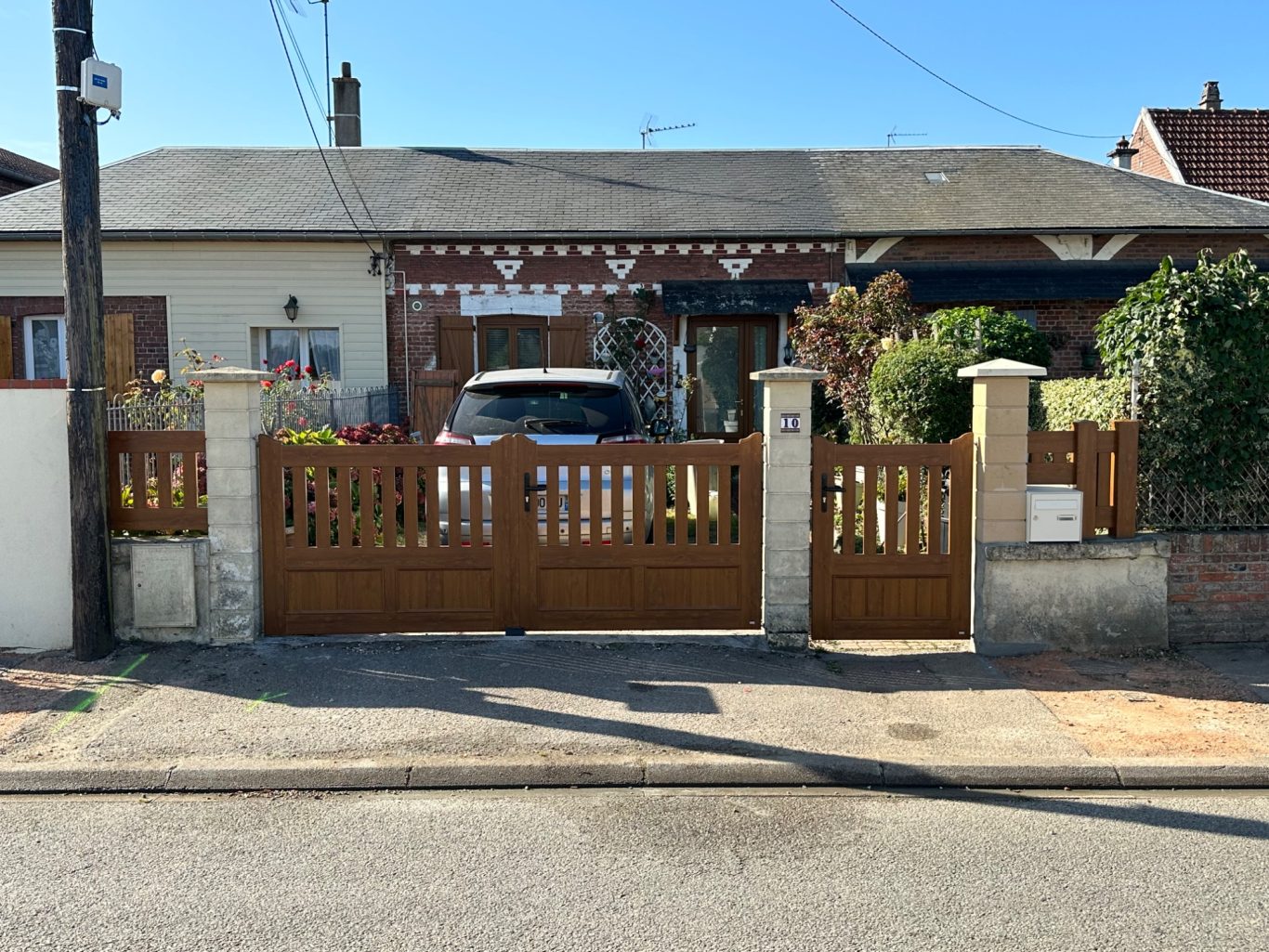 Maison à un étage avec portail en bois et jardin, sous un ciel bleu dégagé.