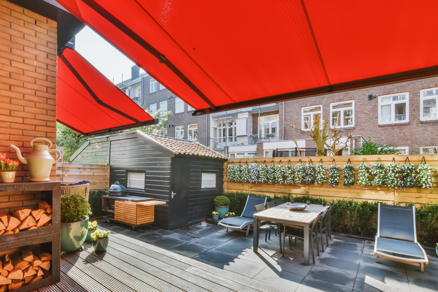 Terrasse ensoleillée avec parasols rouges, table, chaises et murs végétalisés.
